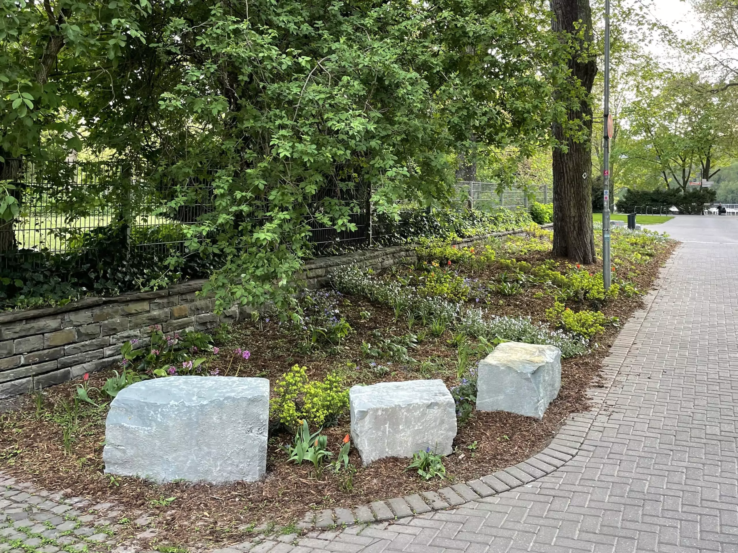 Natursteinquader als Sitzsteine in bepflanzter Grünanlage am Gehweg.