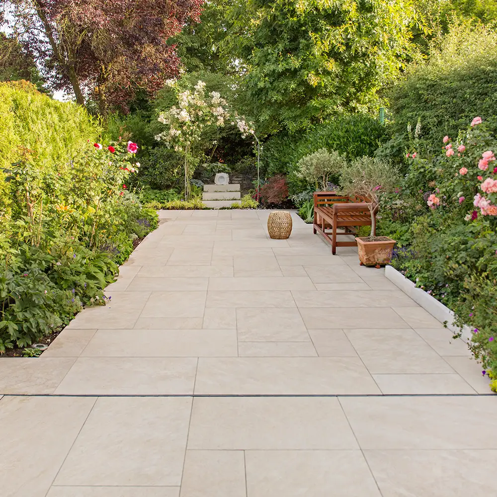 Heller Gartenweg mit Natursteinplatten im Römischen Verband, eingefasst von blühenden Beeten und Gartenmöbeln