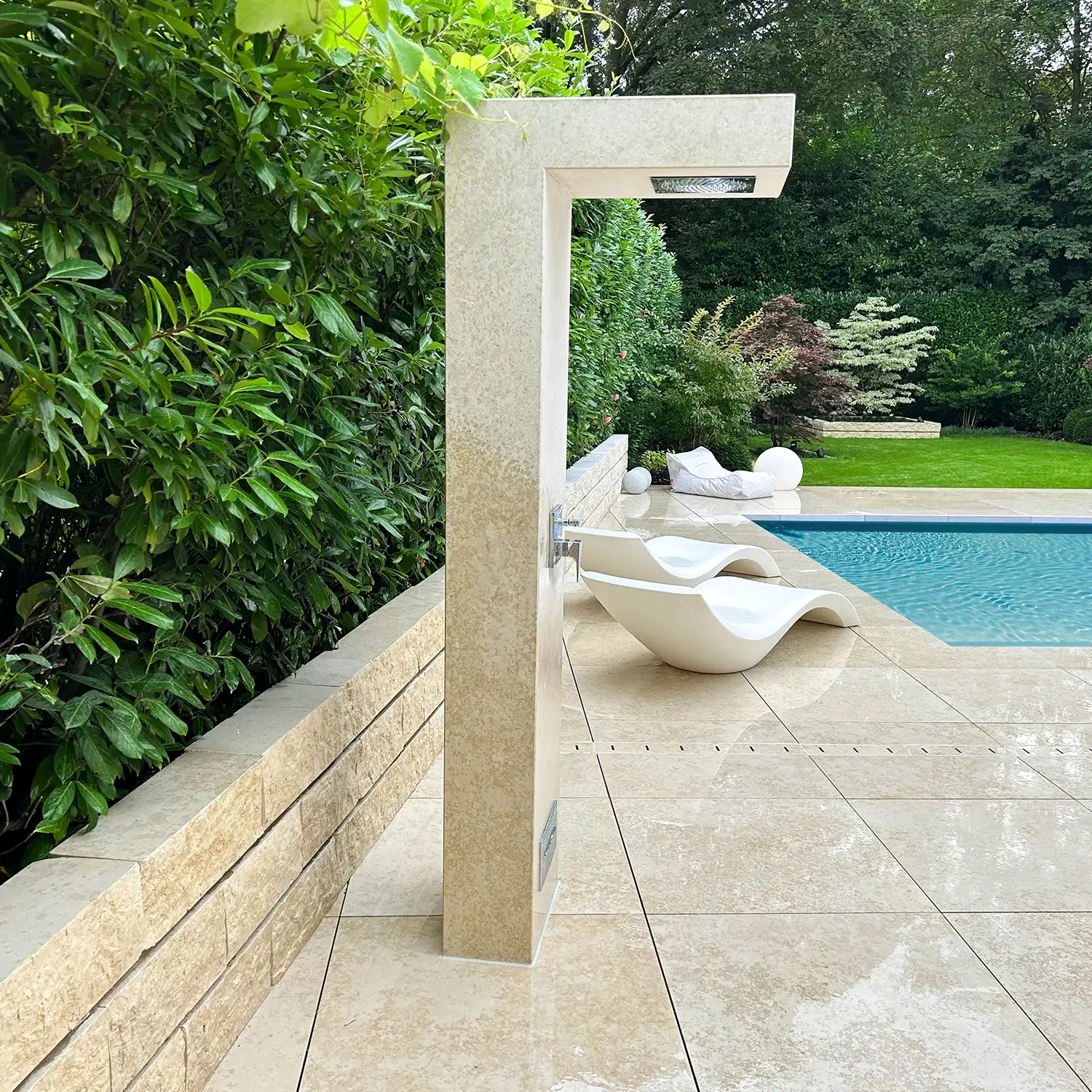 Beige Outdoordusche aus Naturstein Kanfanar neben einem Pool, umgeben von grünen Pflanzen und Gartenmöbeln auf Natursteinplatten
