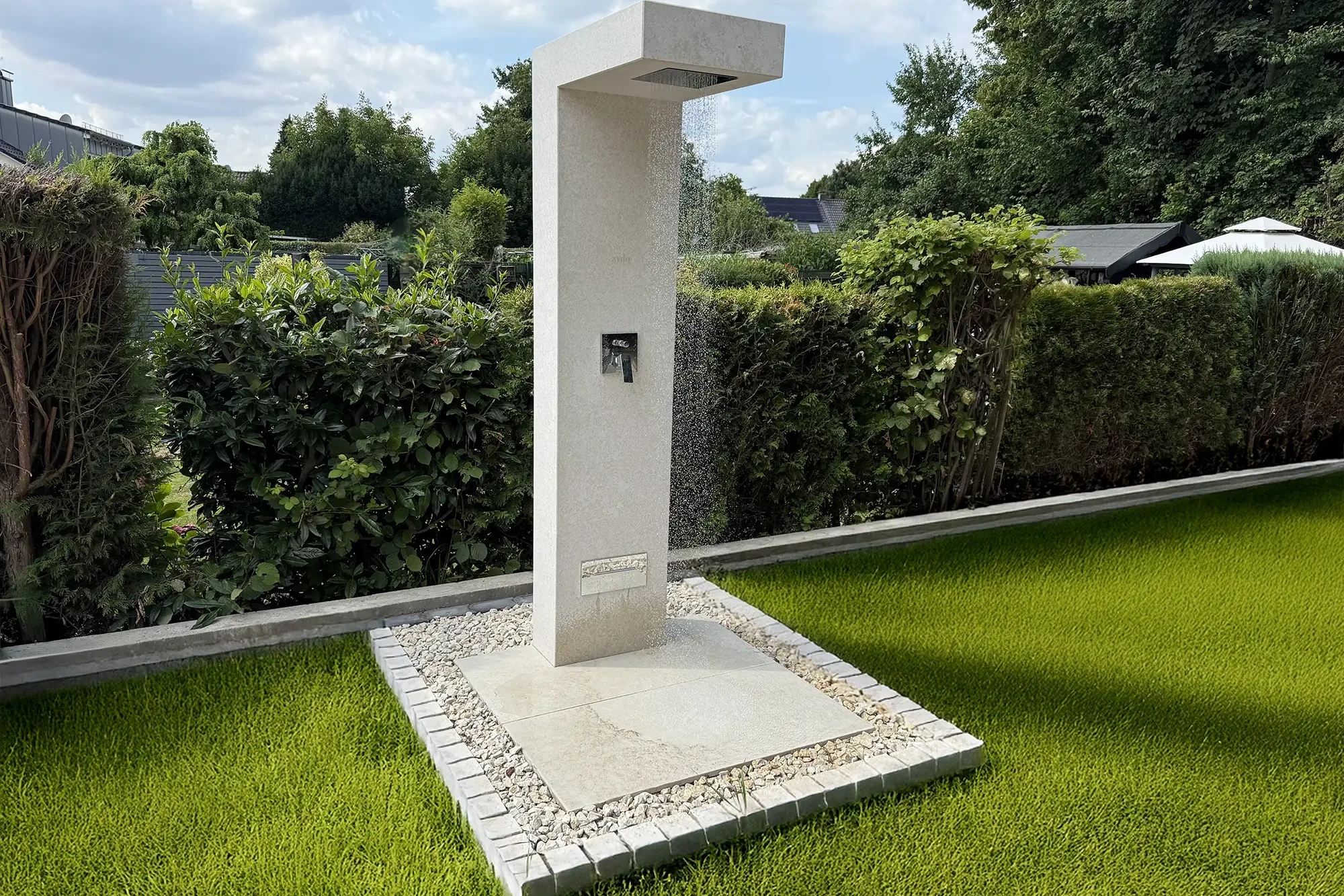 Freistehende Outdoordusche aus hellem Naturstein Kanfanar, montiert auf einer quadratischen Natursteinfläche mit Kiesumrandung, in einem gepflegten Garten mit Hecken und Rasen