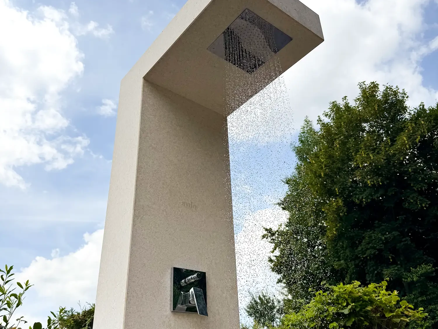 Nahaufnahme einer freistehenden Outdoordusche aus hellem Naturstein Kanfanar mit integrierter Raindance Kopfbrause, aus der Wasserstrahlen nach unten fließen, vor blauem Himmel und Gartenbäumen