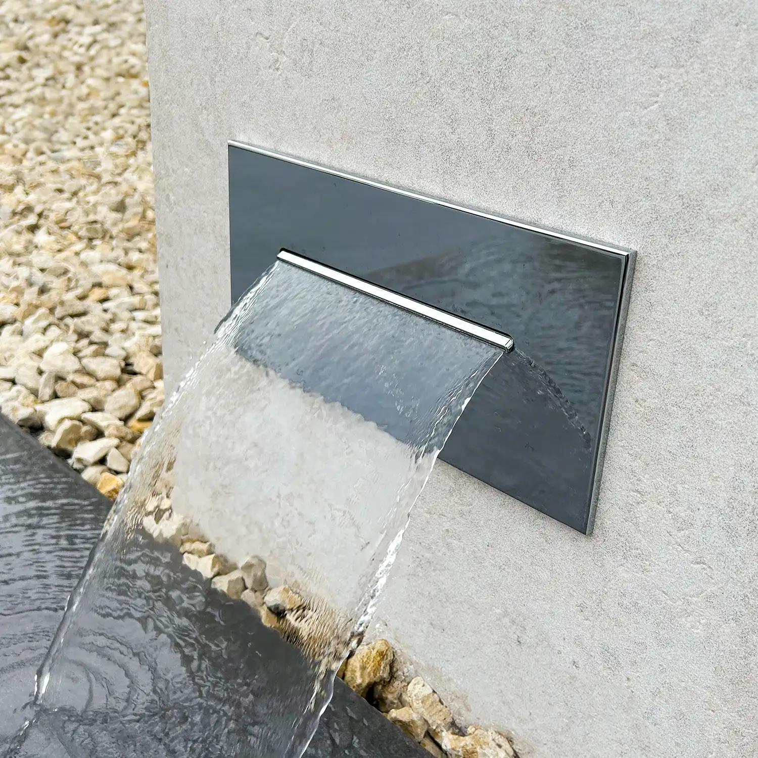 Nahaufnahme einer verchromten Fußdusche mit Wasserfall-Auslass, aus der gleichmäßig Wasser in einen Natursteinbereich fließt