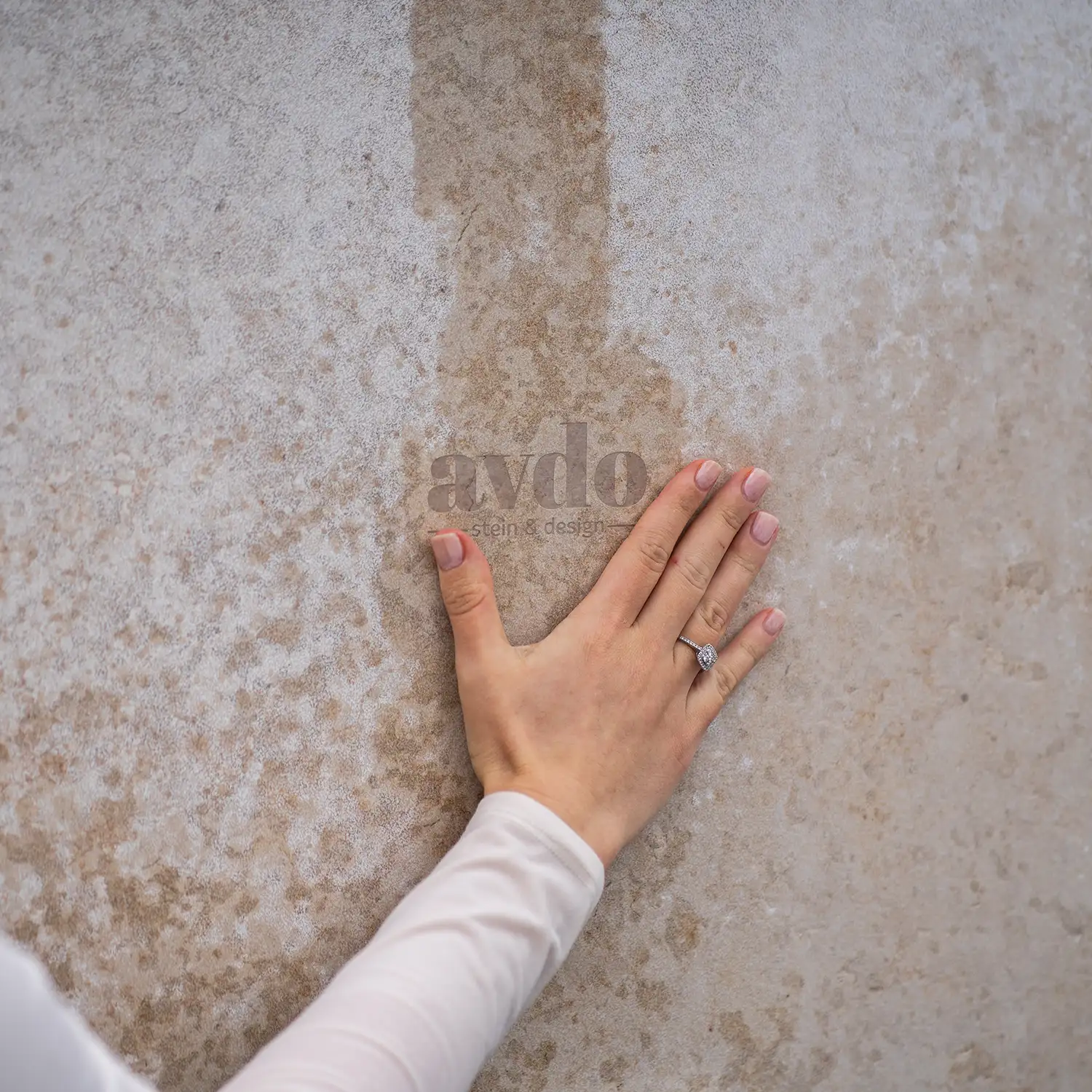 Hand mit Ring berührt die beige-graue, sandgestrahlte Oberfläche des Natursteins Kanfanar und zeigt die natürliche Struktur des Materials