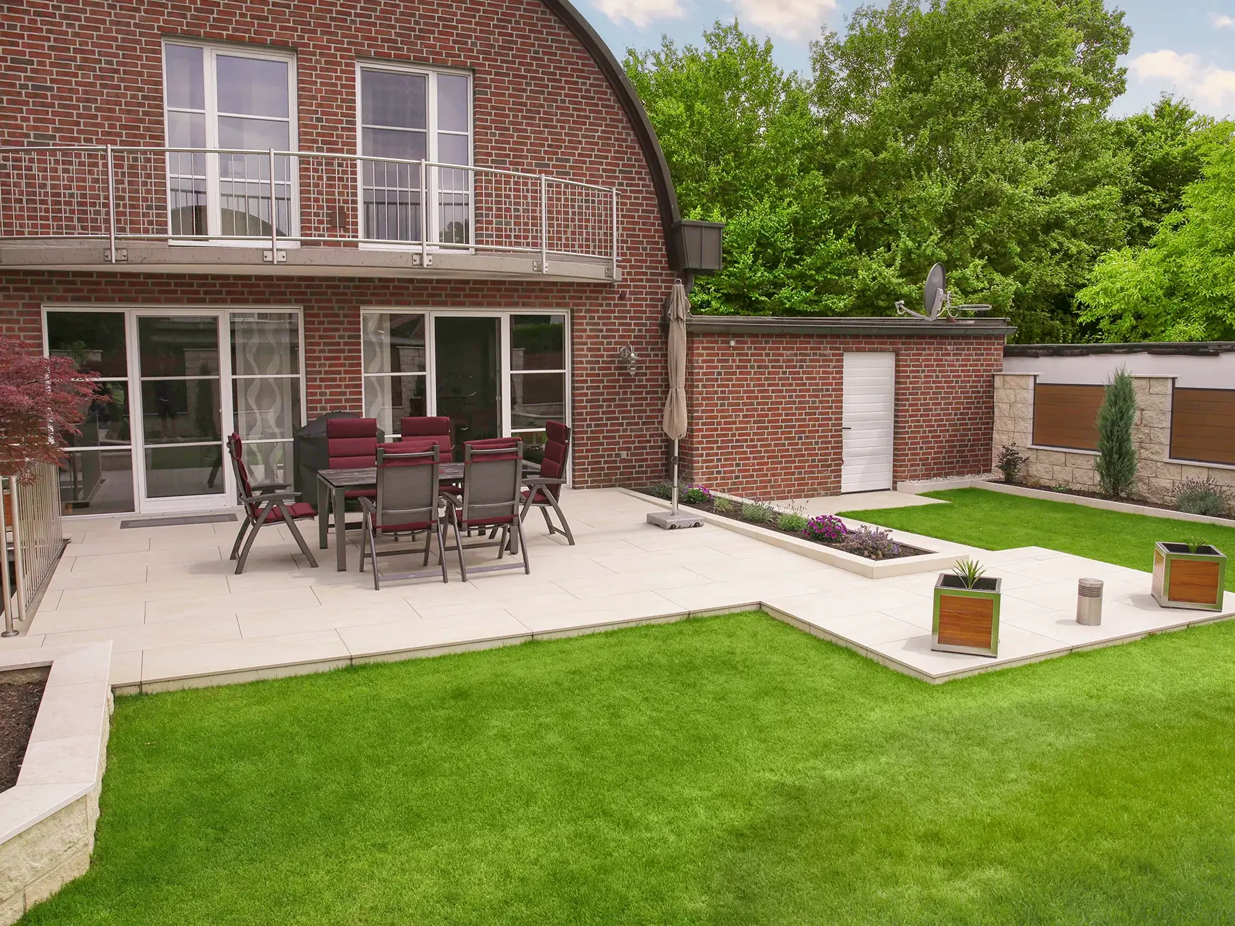 Gepflasterte Terrasse mit cremefarbenen Großformatplatten an Backstein-Einfamilienhaus mit Gartenessbereich, Rasenfläche und Bepflanzung