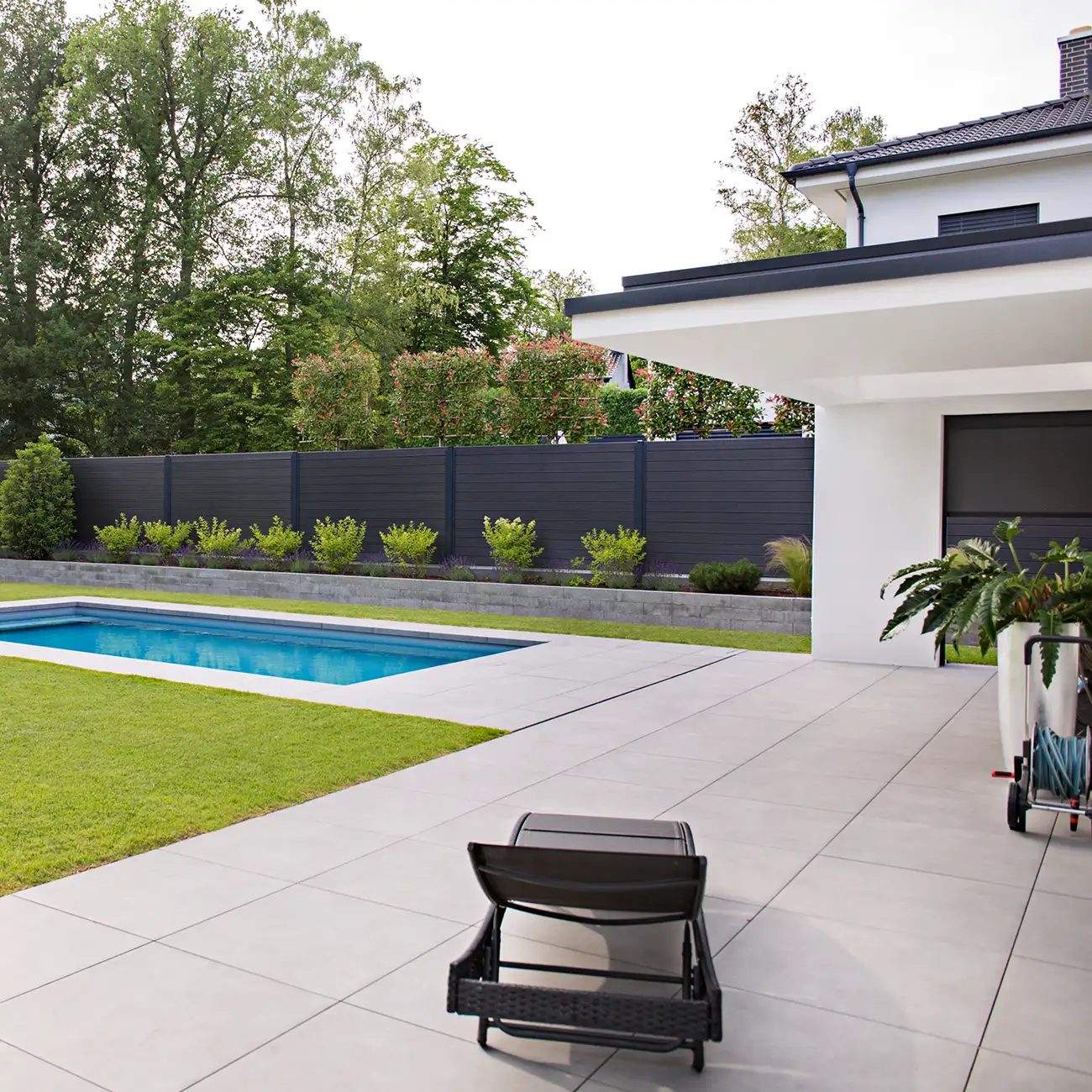 Moderner Au&szlig;enbereich mit gro&szlig;formatigen hellgrauen Keramikplatten, Swimmingpool, Rasenfl&auml;che und wei&szlig;em Einfamilienhaus mit Carport
