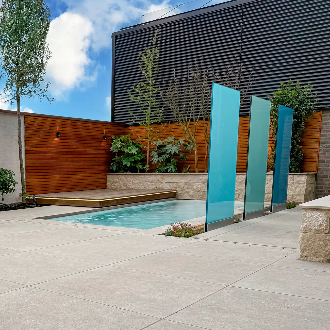 Moderner Designgarten mit hellgrauen Keramikplatten in Betonoptik, kleinem Einbaupool, t&uuml;rkisen Glastrennw&auml;nden, Holzsichtschutz und Holzdeck mit LED-Beleuchtung