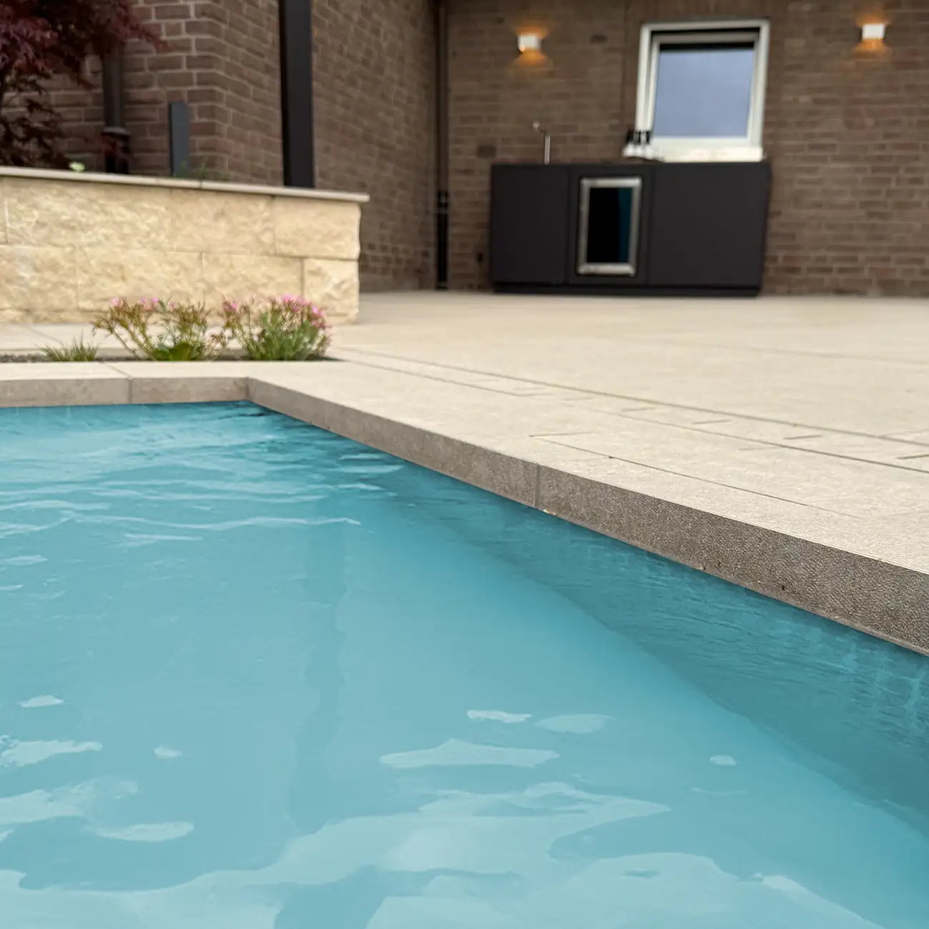Nahaufnahme Poolumrandung aus hellbeigen Keramikplatten mit geflammter Kantenausbildung am t&uuml;rkisblauen Schwimmbecken, Backsteinhaus im Hintergrund