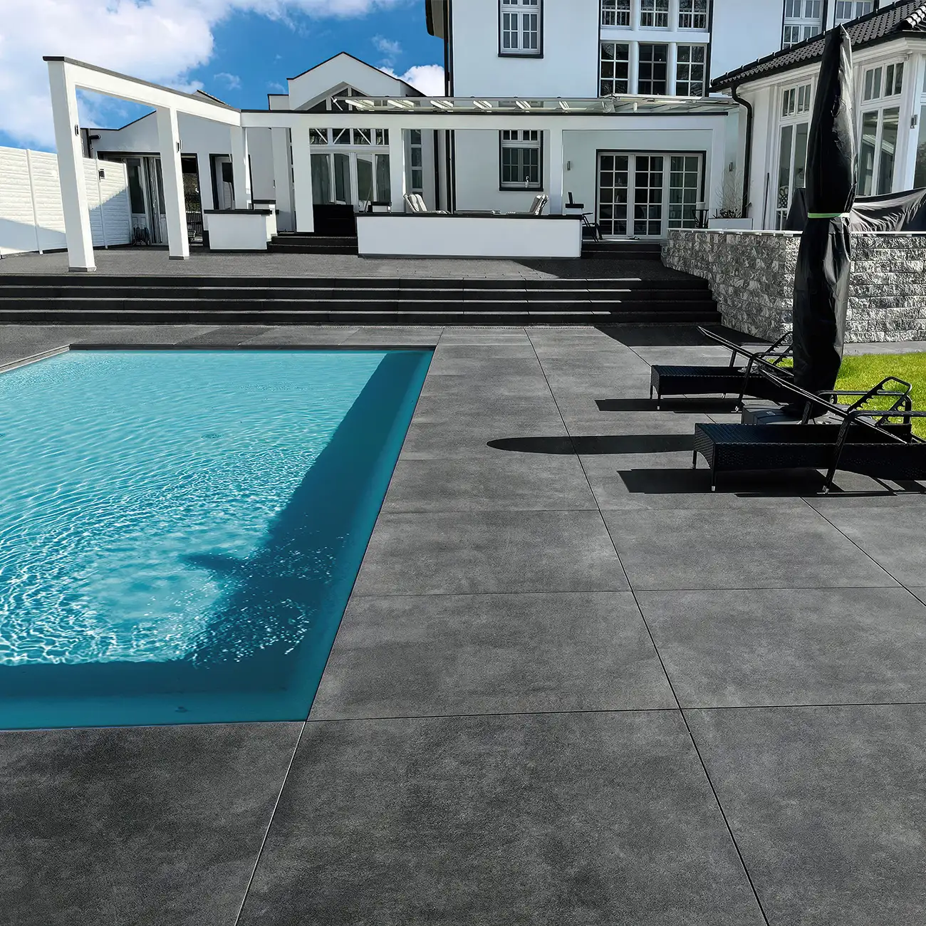 Gro&szlig;z&uuml;gige Poolterrasse mit anthrazitfarbenen Keramikplatten in Betonoptik, breiter Treppenanlage, Liegest&uuml;hlen und wei&szlig;er Villa mit Wintergarten im Hintergrund