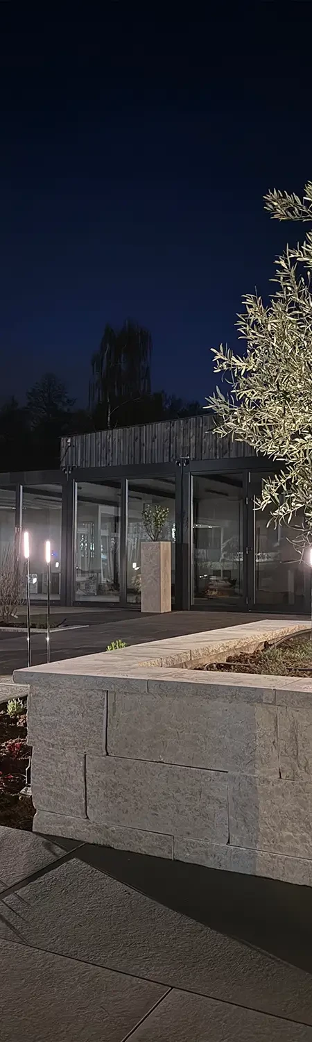 Naturstein-Hochbeet mit Olivenbaum und Gartenbeleuchtung in der Gartenausstellung avdo design M&uuml;lheim an der Ruhr bei Nacht