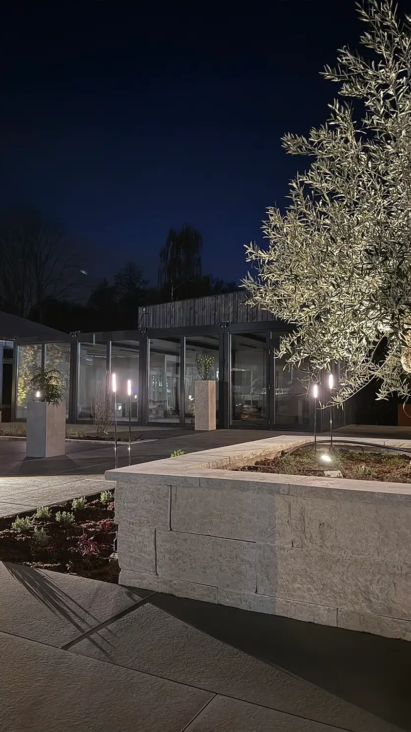 Naturstein-Hochbeet mit Olivenbaum und Gartenbeleuchtung in der Gartenausstellung avdo design M&uuml;lheim an der Ruhr bei Nacht