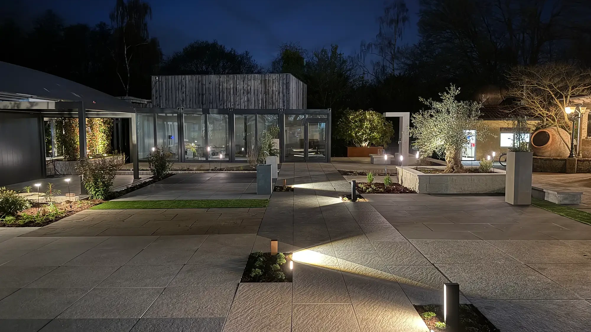 Beleuchtete Naturstein-/ Keramikplatten-Terrasse der Gartenausstellung avdo design in Mülheim an der Ruhr bei Nacht mit Gartenbeleuchtung und Glaspavillon