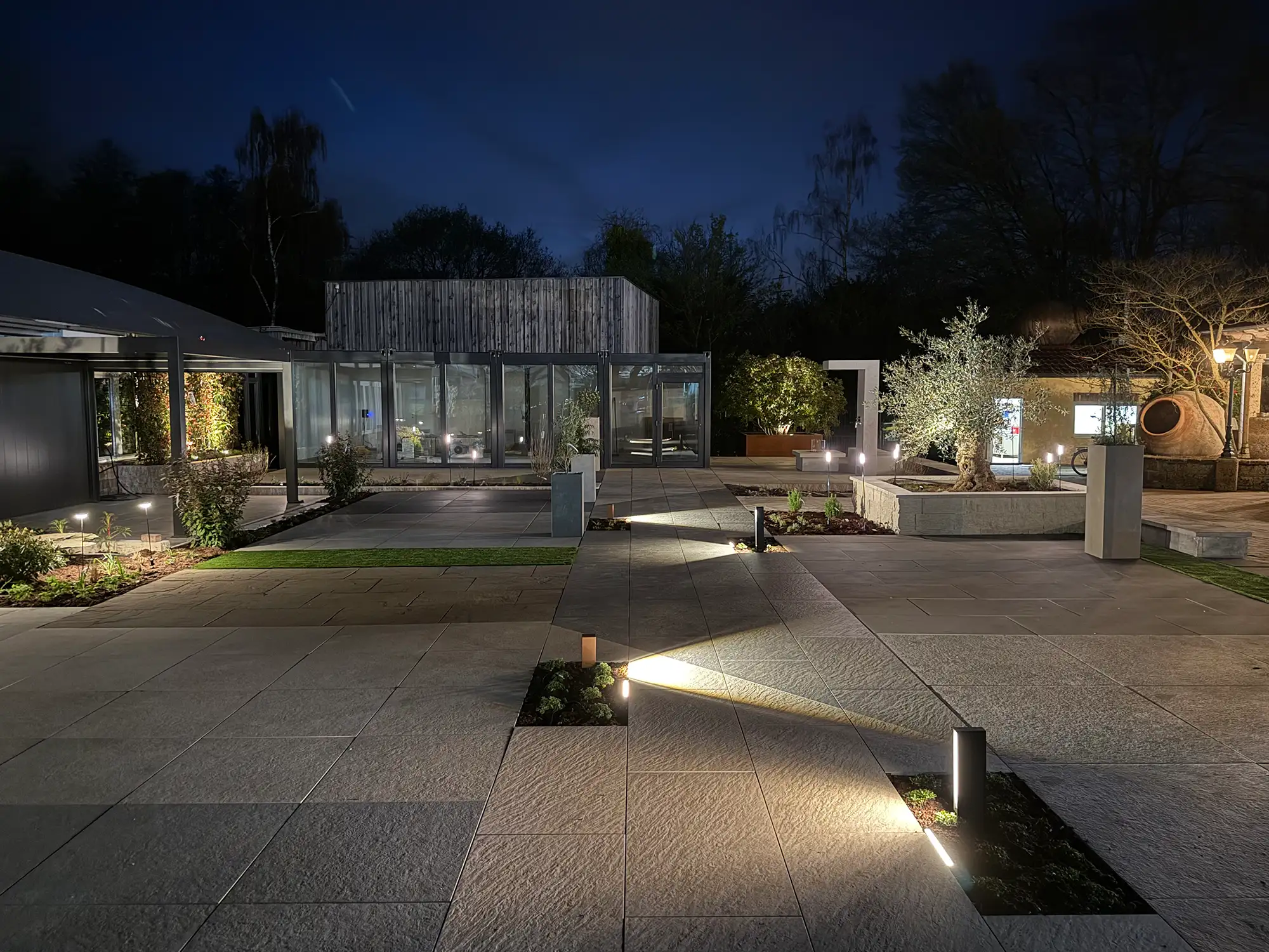 Beleuchtete Naturstein-/ Keramikplatten-Terrasse der Gartenausstellung avdo design in Mülheim an der Ruhr bei Nacht mit Gartenbeleuchtung und Glaspavillon