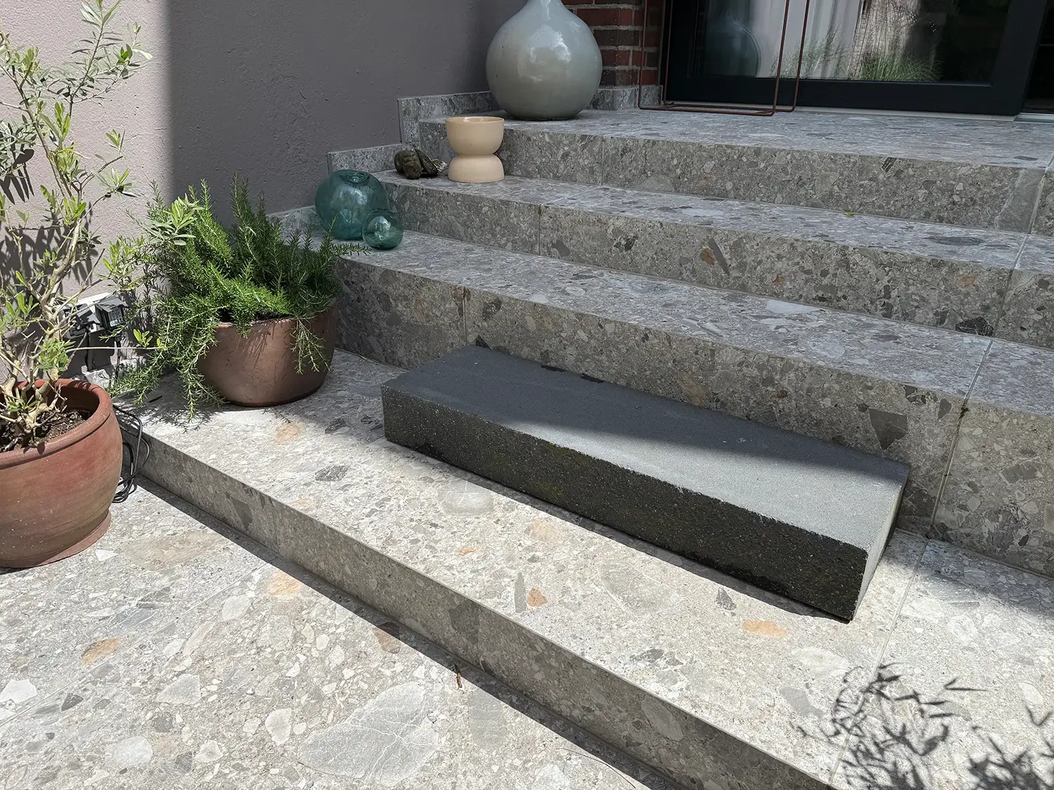 Graue Naturstein-Treppenstufen im Außenbereich mit dunkler Blockstufe, Pflanzgefäßen und modernem Hauseingang