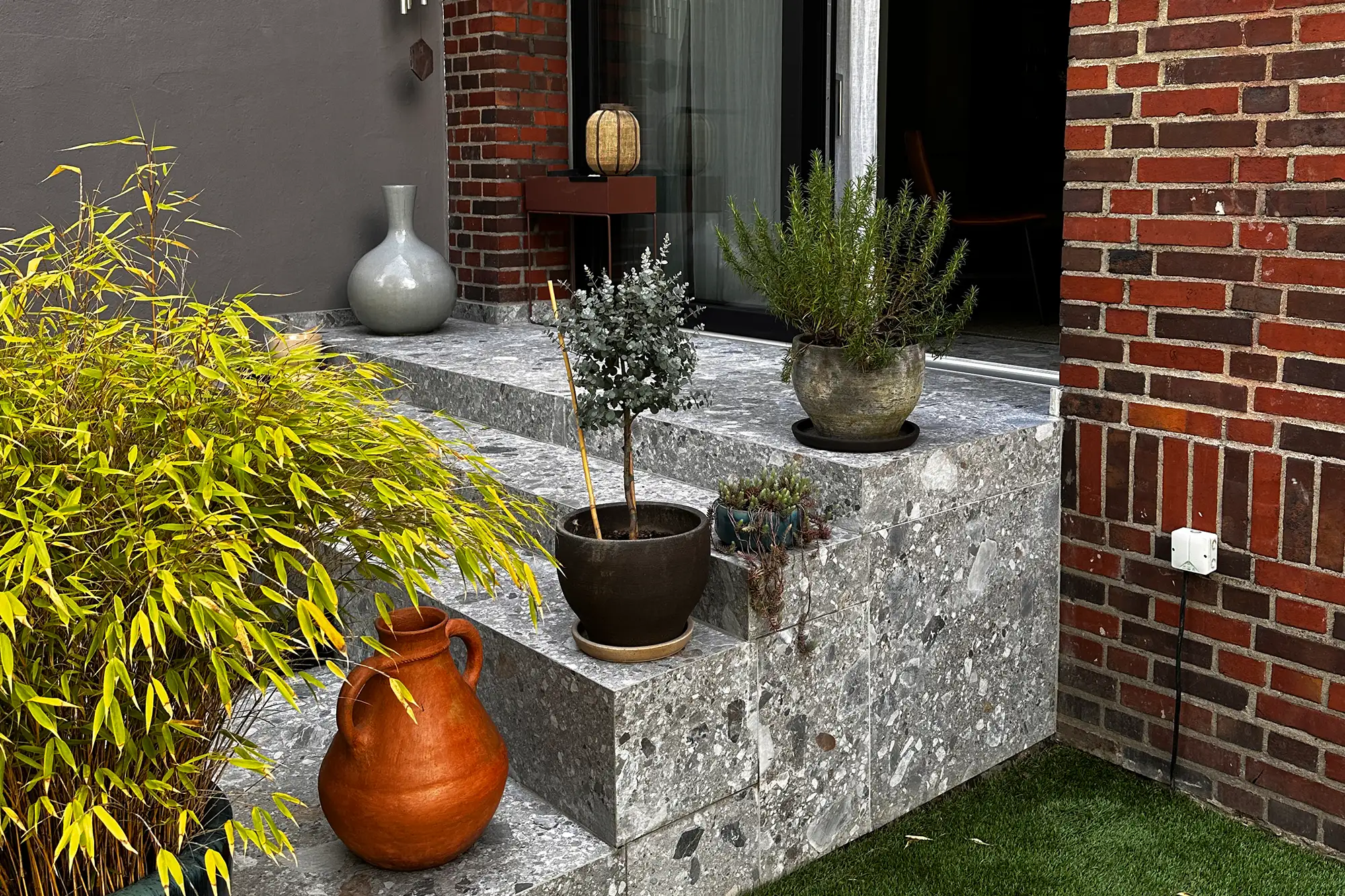 Terrasse mit grauen Steinplatten in Terrazzo-Optik, Stufen, Pflanzgefäßen und dekorativer Bepflanzung am Hauseingang