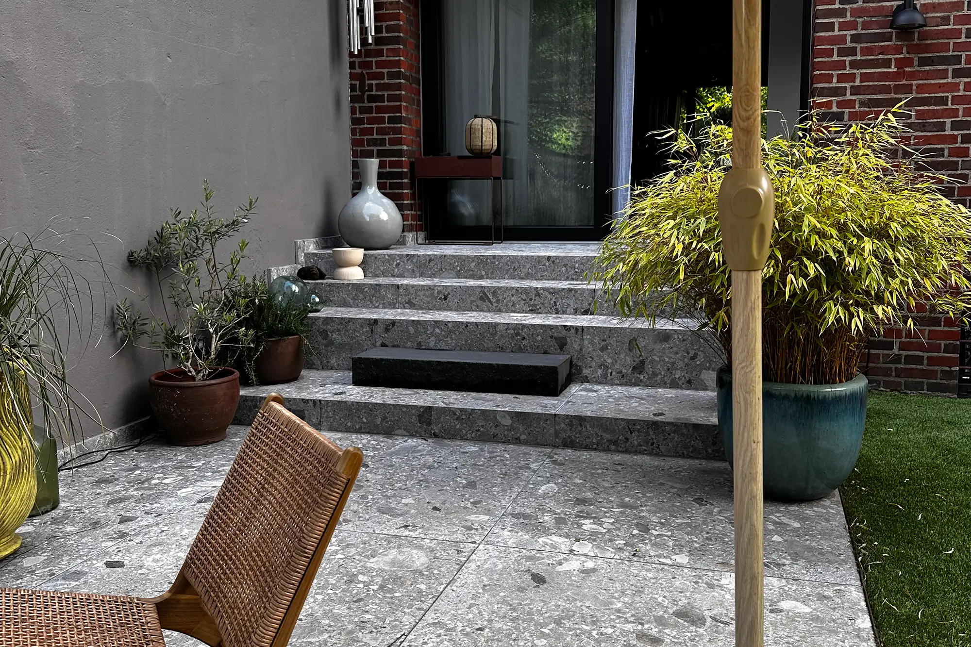 Terrasse mit grauen Steinplatten, Naturstein-Stufen, Rattansessel, Sonnenschirm und dekorativer Bepflanzung