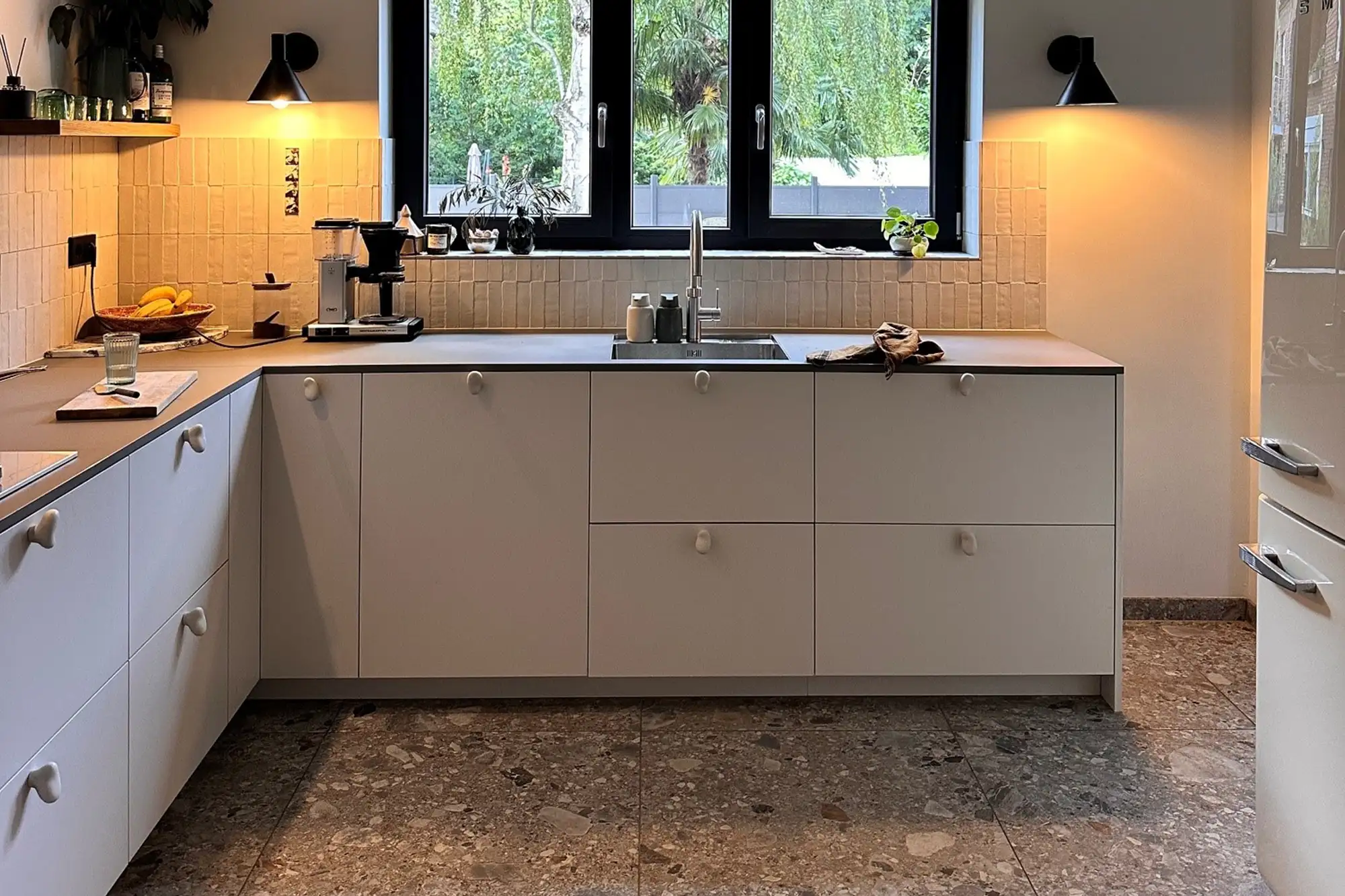 Moderne Küche mit hellem Küchenblock, Steinboden, gefliester Rückwand, Fensterfront und hochwertiger Arbeitsfläche