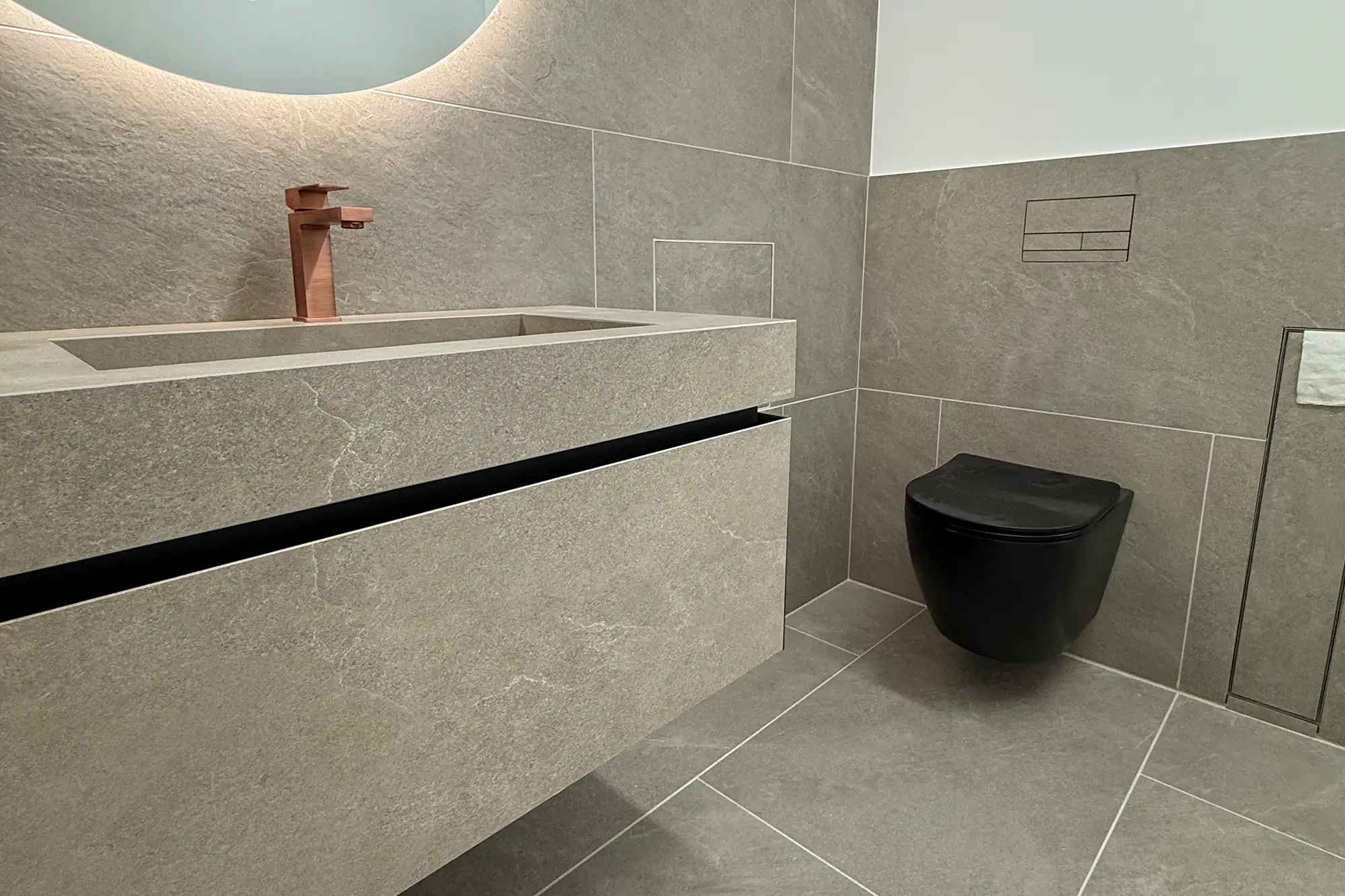 Moderner Waschtisch in grauer Steinoptik mit integriertem Becken, Kupferarmatur, schwarzem WC und hochwertiger Badgestaltung