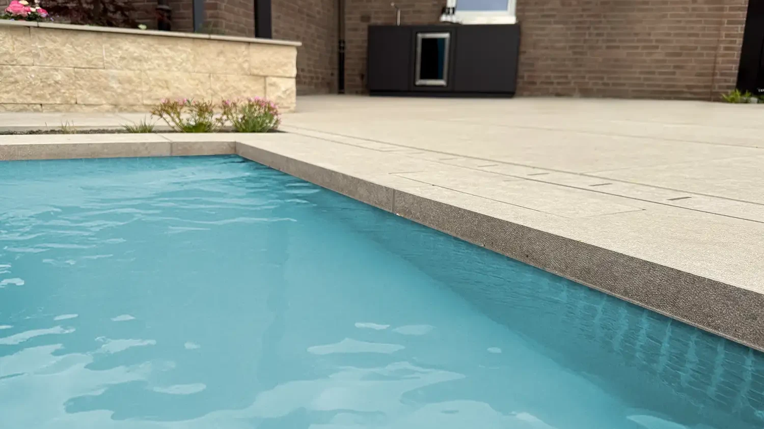 Poolumrandung aus beigen Keramikplatten in Natursteinoptik am Schwimmbecken, Backsteinhaus im Hintergrund.