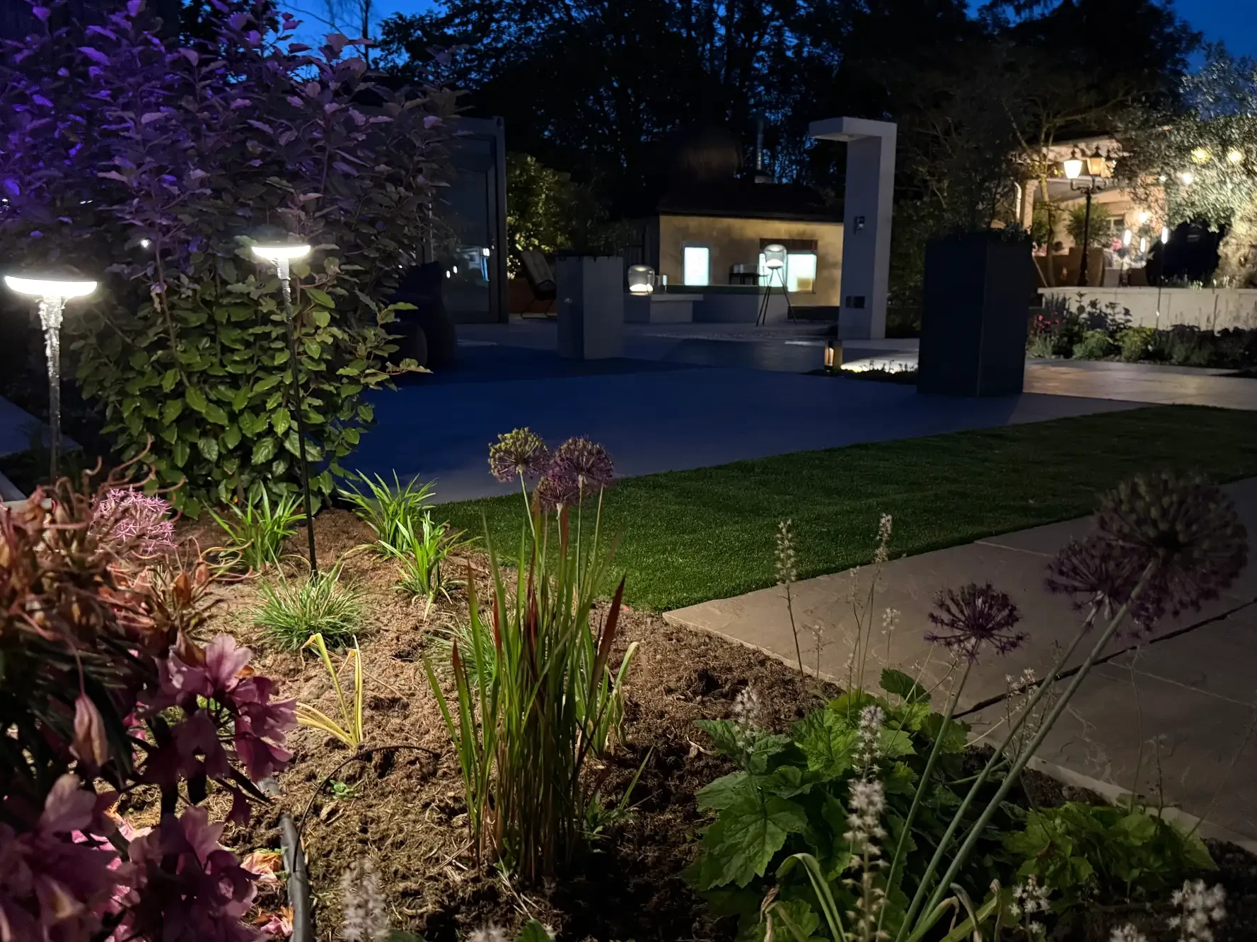 Gartenausstellung von avdo design mit Gartenbeleuchtung, bepflanztem Beet, Rasenfläche und modernen Terrassenplatten bei Nacht