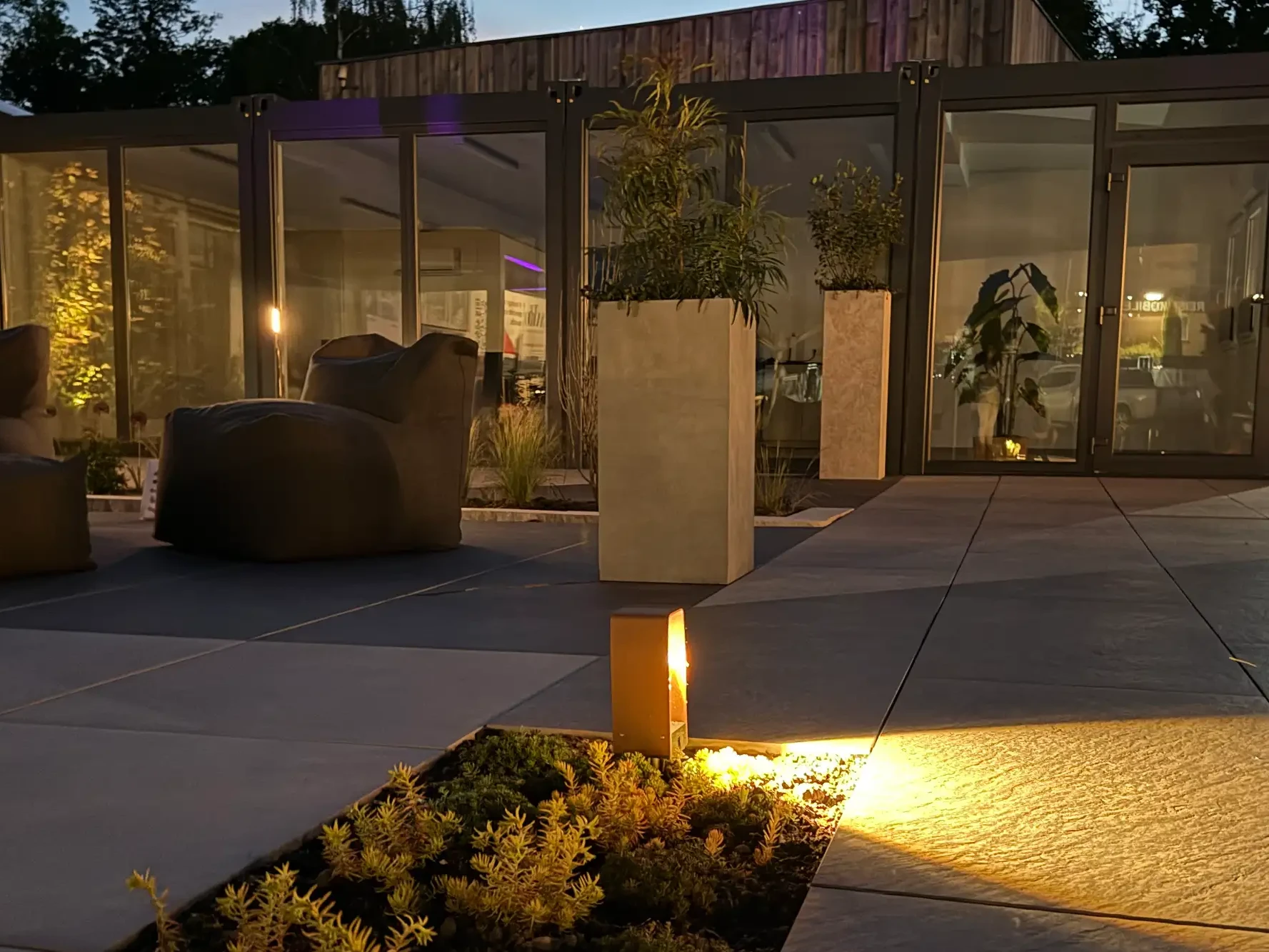 Moderne Gartenausstellung von avdo design mit Keramikplatten, Outdoor-Lounge, Pflanzkübeln, Glasfassade und Gartenbeleuchtung bei Dämmerung