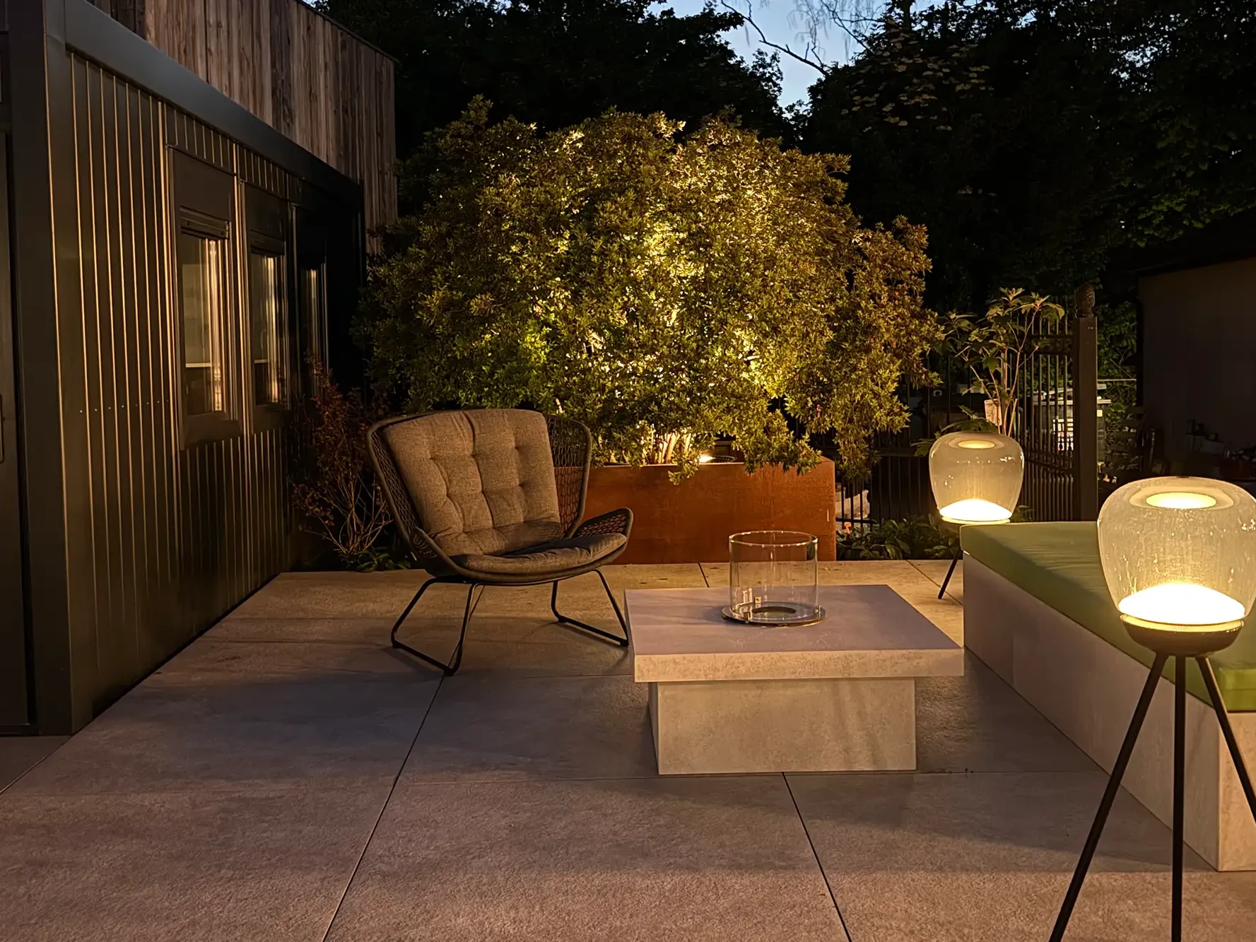 Outdoor-Lounge in der avdo design Gartenausstellung mit modernen Terrassenplatten, Sitzbereich, Gartenbeleuchtung und bepflanztem Außenbereich bei Dämmerung