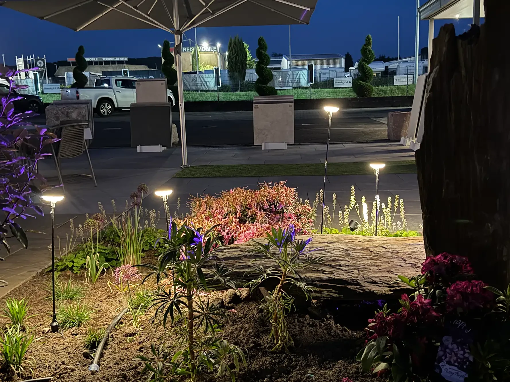 Beleuchteter Natursteinbereich in der avdo design Gartenausstellung mit Pflanzenbeet, Gartenleuchten und Terrassenplatten bei Nacht