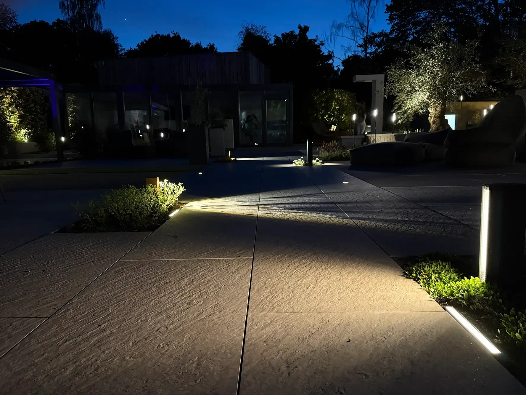 Beleuchtete Terrassenplatten in der avdo design Gartenausstellung mit Gartenleuchten, Pflanzenbeeten und moderner Außenbeleuchtung bei Nacht