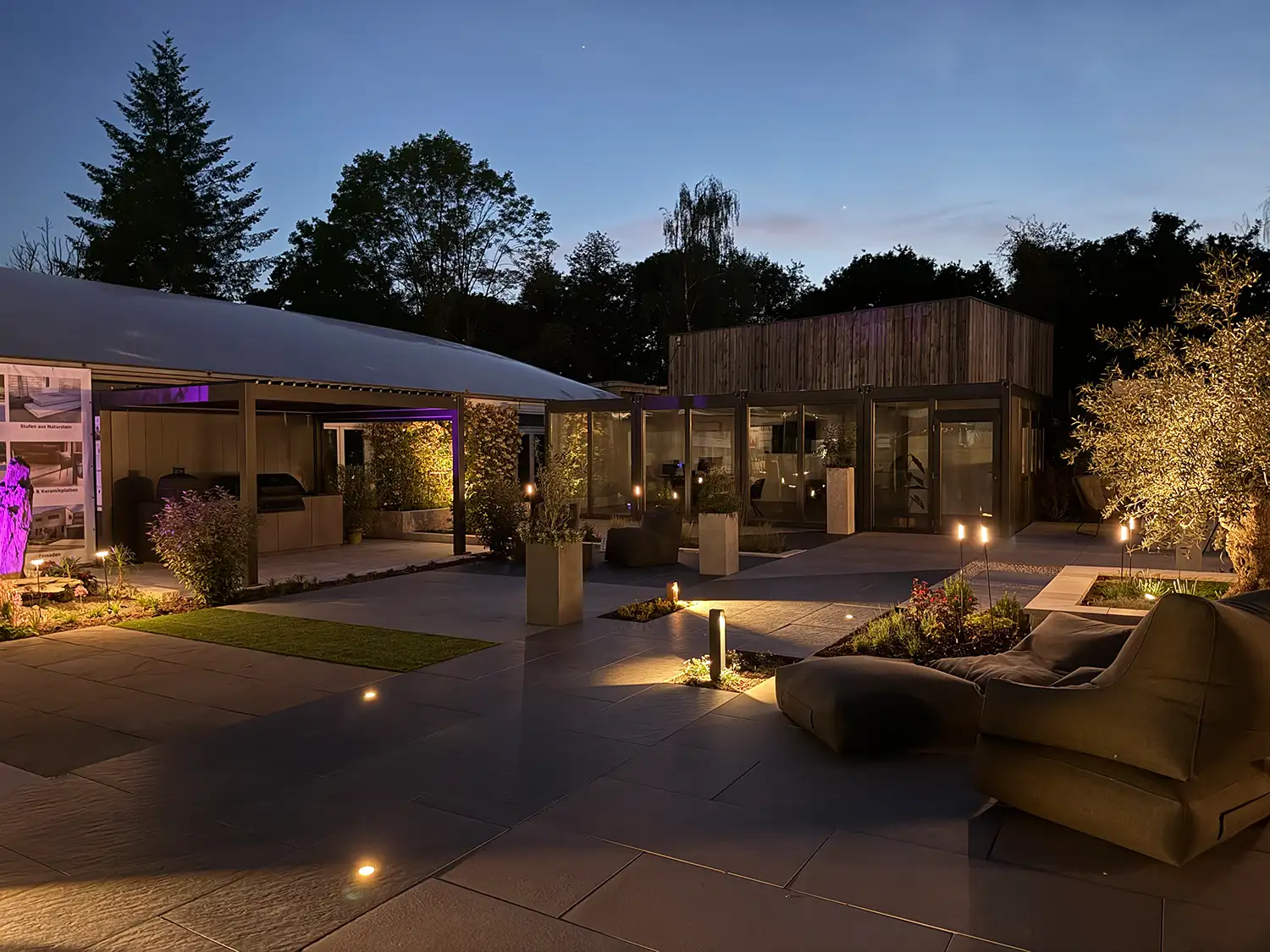 Gartenausstellung von avdo design bei Dämmerung mit Natursteinplatten, Keramik-Terrasse, Outdoor-Lounge und moderner Gartenbeleuchtung