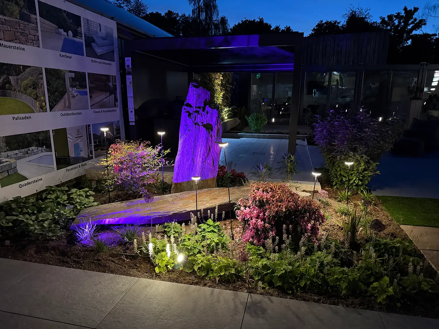 Gartenausstellung von avdo design mit beleuchtetem Naturstein, bepflanztem Gartenbereich, Terrassenplatten und moderner Außenbeleuchtung bei Nacht