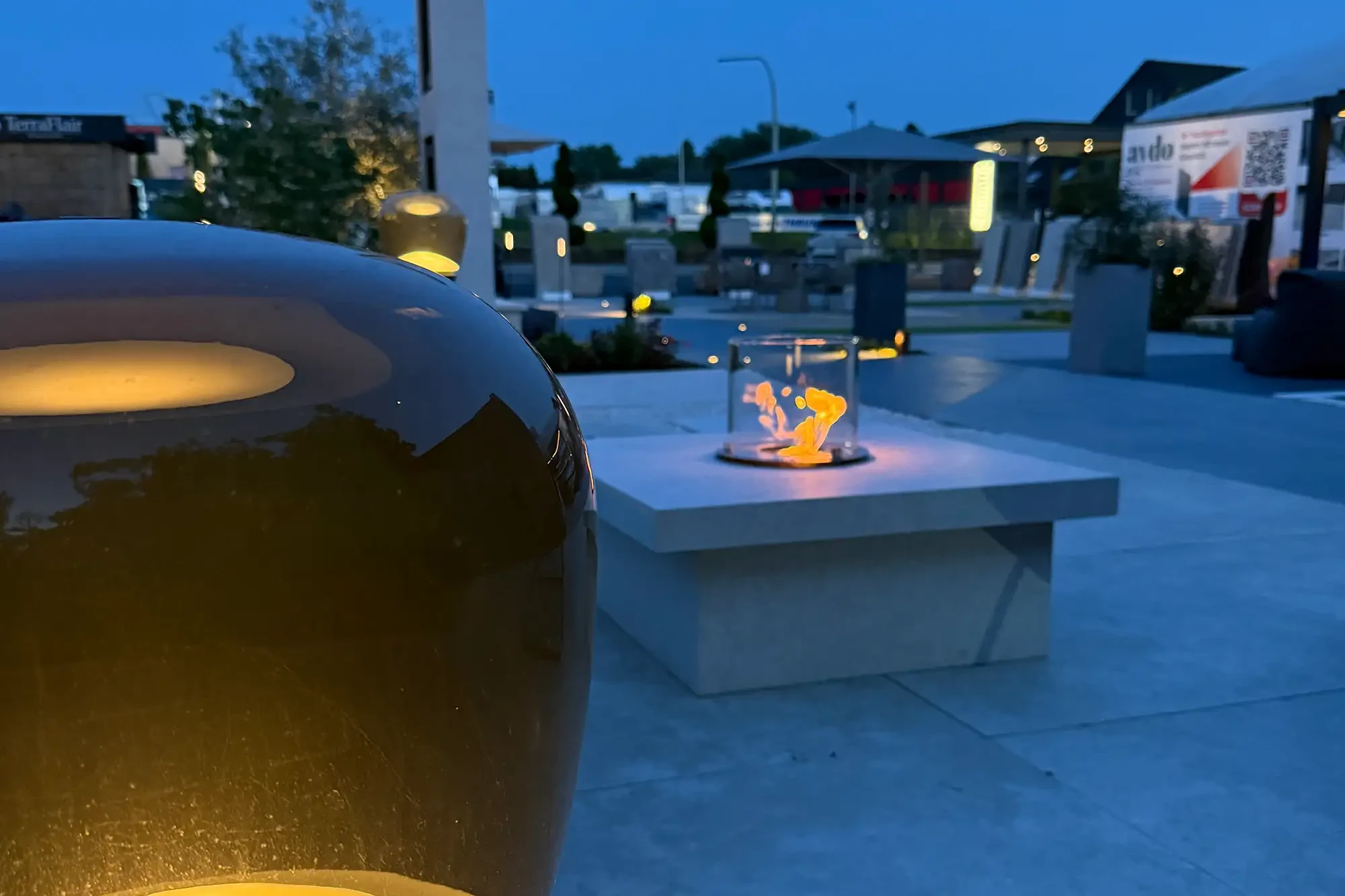 Kamintisch mit Glas-Windschutz und Flamme in moderner Gartenausstellung bei Abendlicht.
