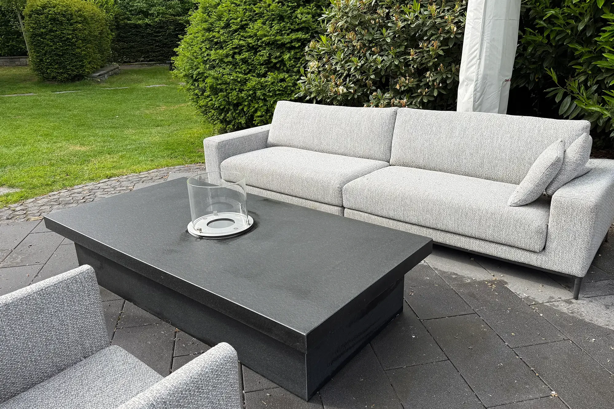 Dunkler Kamintisch mit Glas-Windschutz vor grauer Gartenlounge im Außenbereich.